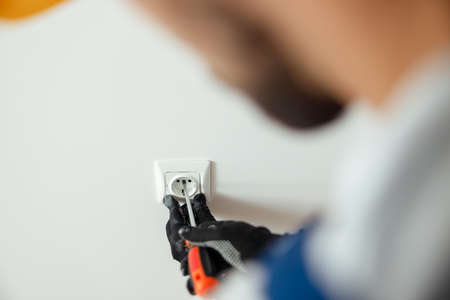Close up of hands of male worker, professional electrician in protective gloves using screwdriver while installing new electrical socket outlet in a roomの写真素材