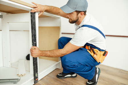 Professional handyman, male worker in uniform using spirit level while checking measurements of kitchen cabinet indoorsの写真素材