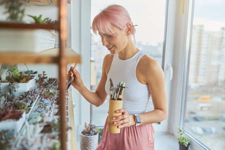 Pretty pink haired woman takes care of houseplants on balcony at homeの写真素材