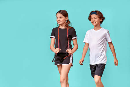 Two happy teenagers, boy and girl with jumping rope looking away and smiling while standing isolated over blue background, exercising togetherの写真素材