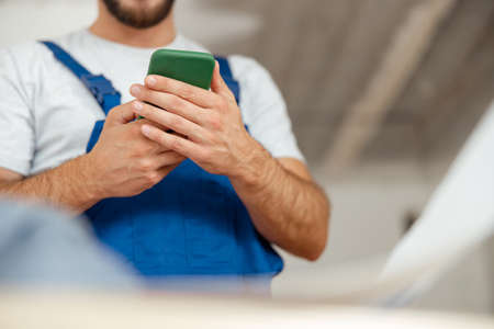 Closeup of hands of repairman in workwear texting, using smartphone, standing indoors during renovation workの写真素材