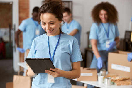 Portrait of charming young female volunteer in blue uniform using tablet pc and smiling while standing indoors. Team sorting, packing items in the backgroundの写真素材