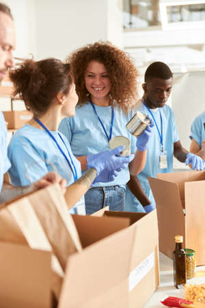 Cheerful volunteers in protective gloves sorting, packing canned food in cardboard boxes, working together on donation project indoorsの写真素材