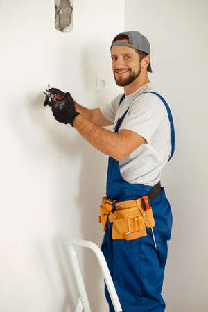 Happy handyman electrician smiling at camera, holding pliers while working on wiring in new houseの写真素材