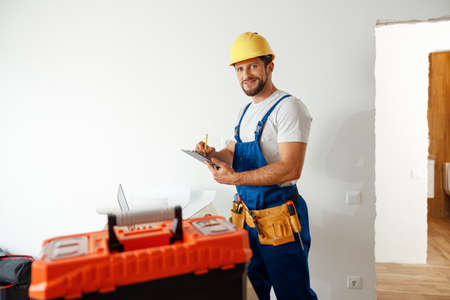 Smiling handyman worker, builder in uniform and hard hat looking at camera, making notes on a clipboard while standing indoorsの写真素材