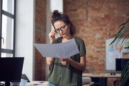 Serious creative woman, interior designer or architect wearing glasses looking focused while working on a blueprint for new project, standing in her officeの写真素材