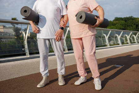 Mature couple in sportswear with mats ready for workout on footbridge at sunset closeupの写真素材