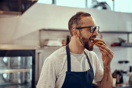 Bearded man in apron eating pastry in cafeの写真素材