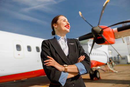 Cheerful woman stewardess standing near plane at airportの写真素材