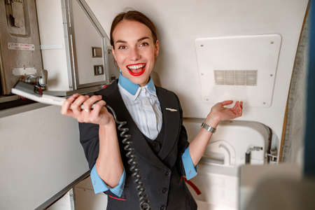 Joyful flight attendant with telephone standing in airplane cabinの写真素材