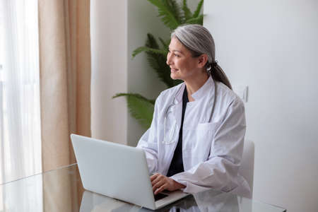 Portrait of happy mature female physician working on laptop in officeの写真素材