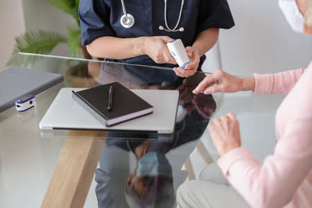 Doctor sitting at the table and holding electronic thermometer, checking temperature of elderly woman in pink sweaterの写真素材