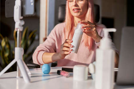 Pretty woman beauty blogger with pink hair shows bottle of cosmetic product at tableの写真素材