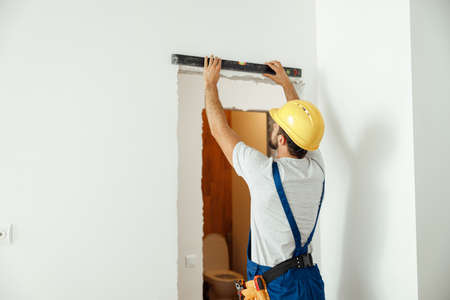 Professional handyman, male worker in uniform using spirit level while taking measurements indoors. Manual work, repair service, occupation conceptの写真素材