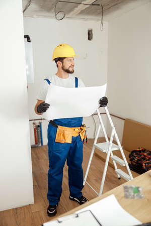 Full length shot of thoughtful contractor in overalls and hard hat looking away while studying blueprint, standing in new apartmentの写真素材