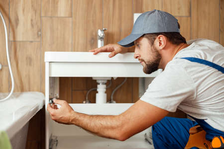Concentrated plumber, male worker in uniform fixing sink and water pipe in the bathroomの写真素材