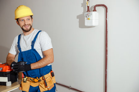 Joyful electrician, handyman in uniform, hard hat and protective gloves smiling aside while posing indoors, leaning on a toolbox. Manual work, maintenance, occupation conceptの写真素材