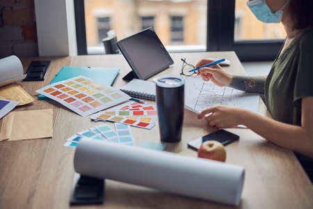 Close up shot of interior designer wearing protective mask working with color samples and blueprint, sitting at the table in her office. Business, Covid19, renovation. Selective focusの写真素材