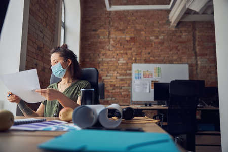 Female interior designer wearing protective mask holding a blueprint while working on new poject, sitting at the table in her office. Business, Covid19, renovation conceptの写真素材
