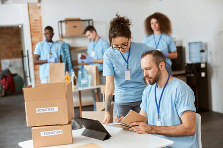 Team of diverse volunteers in blue uniform making notes, calculating, sorting and packing items in cardboard boxes while working together on donation project indoorsの写真素材