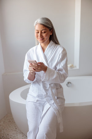 Vertical photo woman sitting on the edge of bathtub using smartphoneの写真素材