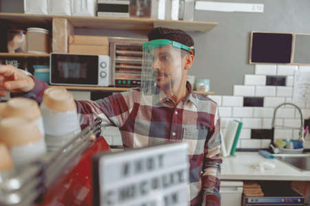 Attractive barista in protective visor standing near coffee machine taking cupの写真素材