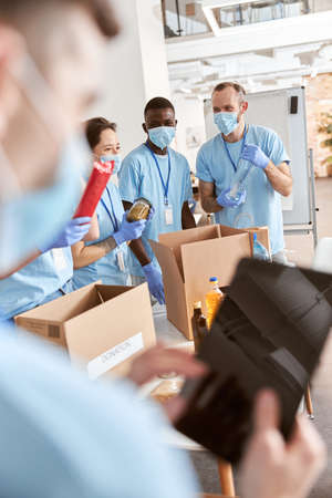 Cheerful volunteers in blue uniform, protective masks and gloves sorting, packing food stuff in cardboard boxes, working together on donation projectの写真素材