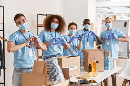 Group of diverse people wearing blue uniform, protective masks and gloves showing love heart sign while volunteering in community, sorting donated food itemsの写真素材