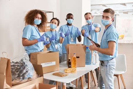 Group of diverse people wearing blue uniform, protective masks and gloves showing thumbs up while sorting donated food items, volunteering in community togetherの写真素材