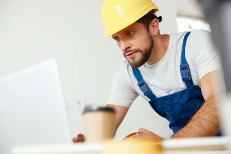 Focused handyman in uniform and hard hat using laptop while starting renovation work indoorsの写真素材