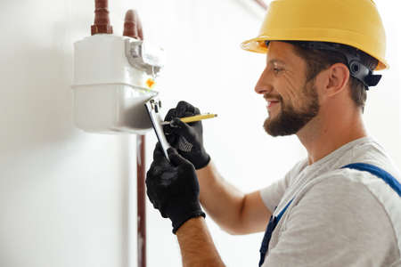 Happy professional heating fitter making notes while checking gas consumption at gas pipe and meter counterの素材 [FY310182332462]