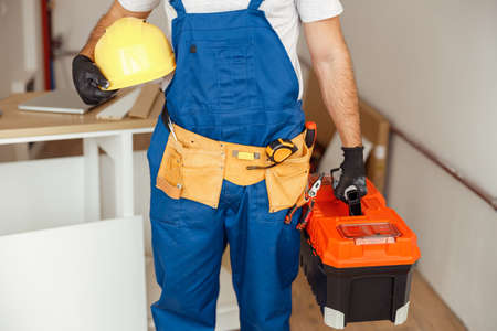 Cropped shot of contractor worker in overalls and hardhat with toolbox standing indoorsの写真素材