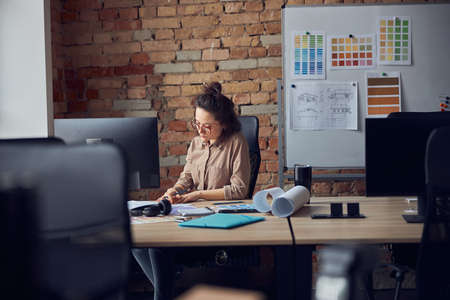 Professional female interior designer looking at blueprint while working on new design project, sitting at the table in her officeの写真素材