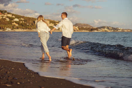 Back photo of happy couple running along the seashore at sunsetの写真素材
