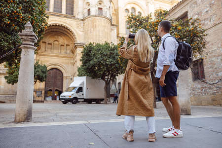 Full-length photo of man and woman tourists taking photo of building while walkingの写真素材