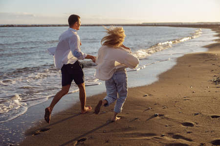 Back photo of man and woman running holding hands along the oceanの写真素材