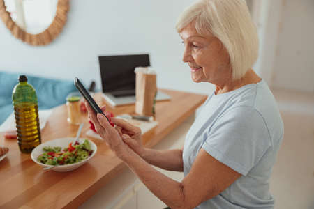 Cheerful pensioner female using mobile phone in kitchenの写真素材