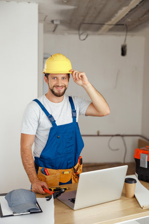 Smiling handyman worker in uniform wearing tool belt and hardhat looking at camera during renovation work indoorsの写真素材