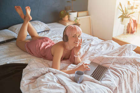 Pink haired woman in silk pajamas rests using laptop and headphones on large bedの写真素材