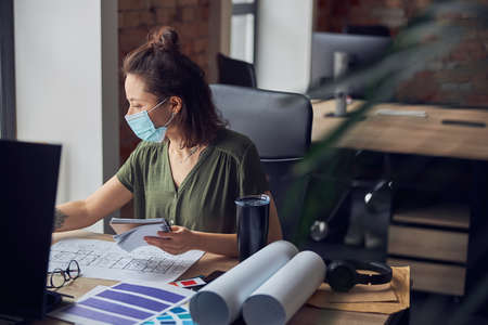 Woman interior designer wearing protective mask making notes in her notebook while working on new project, sitting at the table in her officeの写真素材