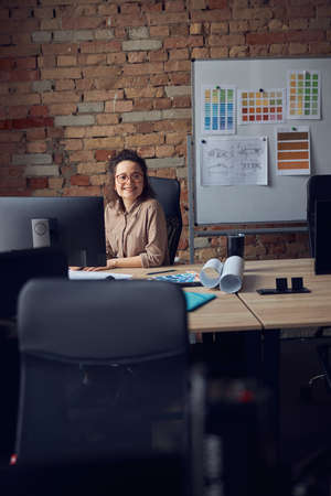 Professional female interior designer in glasses smiling at camera while working on new design project, sitting at the table in her officeの写真素材
