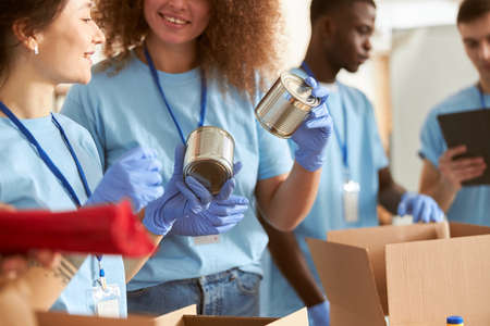 Close up shot of canned food in hands of volunteers in protective gloves sorting, packing foodstuff in cardboard boxes, working together on donation project indoorsの写真素材