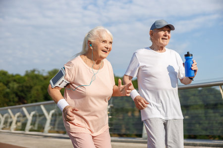 Senior gentleman with cap and bottle and woman with gadgets run along footbridgeの写真素材