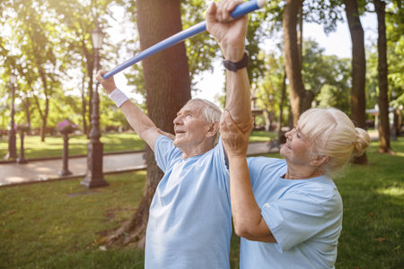 Careful woman helps senior man to do exercise with bar in parkの写真素材