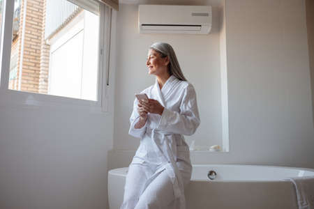 Smiling Asian lady in the bathroom sitting on bathtub and typing on smartphoneの写真素材