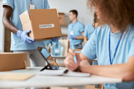 Young female volunteer in blue uniform making notes while working on donation project. Team sorting, packing items in cardboard box in the backgroundの写真素材