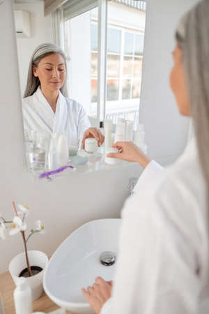 Asian woman in white bathrobe in washroom near sinkの写真素材