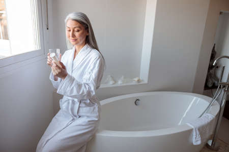 Asian woman sitting on bathtub in bathroom holding cosmetic jarの写真素材