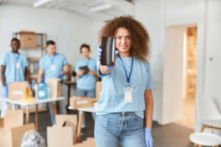 Young woman, volunteer in blue uniform holding, showing smartphone to camera while standing indoors. Team sorting, packing items in the backgroundの写真素材