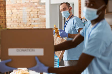 Portrait of male volunteer in blue uniform, protective mask and gloves looking at camera while sorting items. Blurred image of people carrying cardboard box with donated food stuffsの写真素材